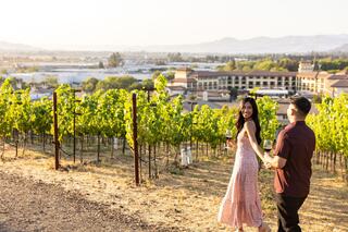 A couple enjoys a romantic moment in a vineyard, holding glasses of wine and taking in the scenic view of lush greenery and distant hills.