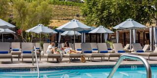 A couple relaxes by a pool, enjoying drinks and snacks under umbrellas, with a backdrop of vineyards and sunlit trees.