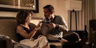 A couple toasts with glasses of red wine in a cozy, warmly lit room, sharing an intimate moment.