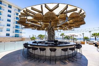 A stylish outdoor bar features a unique umbrella structure with hanging lights, surrounded by greenery and modern seating.