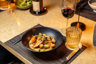 A black bowl filled with shrimp, gnocchi, and greens sits on a stylish table, accompanied by a glass of red wine and a golden drink.