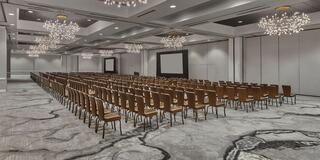 A spacious conference room with rows of brown chairs, elegant chandeliers, and a large screen at the front, set for an event.