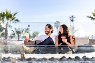 A smiling couple enjoys colorful cocktails on a sunny patio, surrounded by palm trees and a relaxed atmosphere.