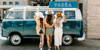 Three stylish women in hats pose together by a vintage blue van, exuding a carefree, summery vibe with a sign reading "Pasea" above.