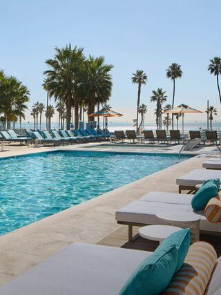 A sunny poolside scene features lounge chairs and umbrellas, palm trees lining the edge, and a serene view of the water.