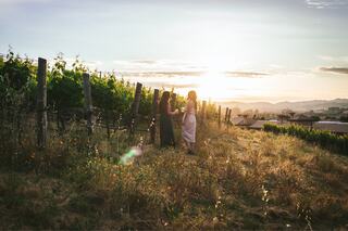 Two women stand in a vineyard at sunset, enjoying their surroundings amidst rows of vines and a backdrop of rolling hills.