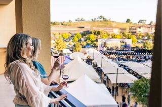 Two women enjoy wine on a balcony overlooking a lively outdoor event with tents, a crowd, and scenic vineyards in the background.