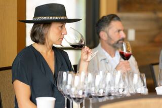 A woman in a hat enjoys wine, thoughtfully swirling her glass, while a man in the background shares the tasting experience.