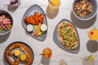 Variety of dishes includes wings with sauce, flatbread with toppings, a pretzel with dipping sauce, and colorful salads, alongside cocktails.