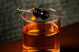 A close-up of a glass filled with amber liquid, garnished with two dark cherries on a skewer, resting on a wooden surface.