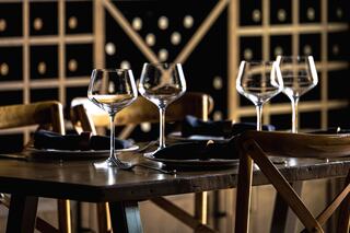A elegantly set table with wine glasses, dark plates, and wooden chairs, complemented by a backdrop of wine storage.