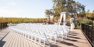 A beautiful outdoor wedding venue features rows of white chairs facing a floral-decorated gazebo, surrounded by lush vineyards under a clear sky.