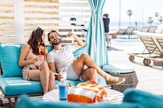 A couple enjoys drinks on a sunny lounge, laughing together amidst a vibrant beachside setting with palm trees in the background.