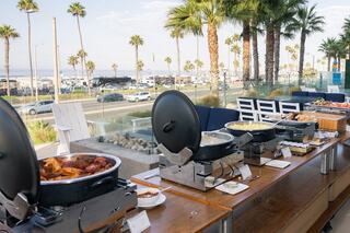 A beachfront buffet features a variety of dishes with palm trees and the ocean in the background, creating a vibrant, inviting atmosphere.