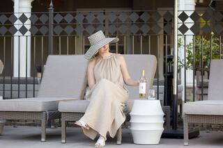 A woman in a stylish hat and beige dress sits on a lounge chair, enjoying a bottle of wine beside a modern white table.