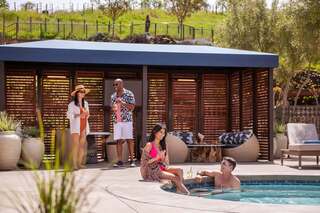 People enjoy a sunny day by the pool, sipping drinks and socializing, surrounded by lush greenery and stylish outdoor furniture.