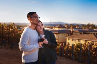 A couple enjoys a romantic moment, with a glass of wine in hand, surrounded by vineyards and a scenic landscape at sunset.