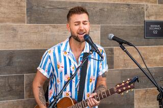 A musician in a colorful, floral shirt passionately sings and plays guitar, surrounded by a wooden wall backdrop and microphones.