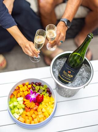 Two people toast with champagne flutes, surrounded by a vibrant fruit platter and an ice bucket holding a bottle of Mumm champagne.