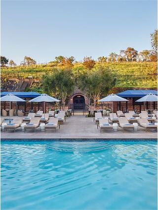 A serene poolside scene with lounge chairs and umbrellas, set against lush greenery and a vineyard backdrop, creating a tranquil retreat atmosphere.