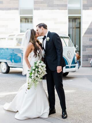 A couple shares a romantic kiss, with the bride in a sleek gown and the groom in a tuxedo, framed by a vintage blue Volkswagen van.