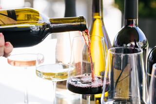 Red wine is being poured into a glass, surrounded by various bottles and glasses of different types of wine in an elegant setting.