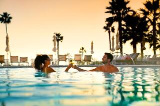 A couple enjoys a toast in a pool at sunset, surrounded by palm trees and lounge chairs, radiating a relaxed and romantic atmosphere.