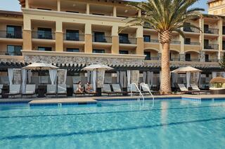 A tranquil resort pool area features lounge chairs under umbrellas, palm trees, and a luxurious building in the background.