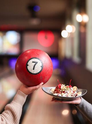 A red bowling ball with the number seven is held in one hand, while another hand offers a plate of ice cream topped with cherries.