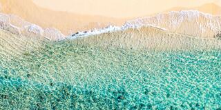 Golden sands meet clear, turquoise waters, with gentle waves lapping at the shore and a solitary figure walking along the beach.