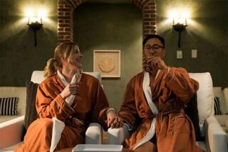 A couple in matching robes enjoys drinks while holding hands, seated comfortably in a cozy, warmly lit space.