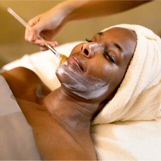 A woman relaxes during a facial treatment, eyes closed, while a hand applies a cream or mask to her face. A tranquil spa atmosphere is evident.