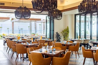 A chic restaurant with warm wooden tables, elegant chandeliers, and plush seating, featuring large windows that overlook a beachside view.