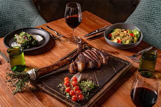 A beautifully arranged dining table features a large grilled steak, fresh salads, cherry tomatoes, and glasses of red wine, creating an elegant setting.
