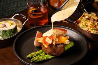 A gourmet dish features a juicy steak topped with lobster, asparagus, and a creamy sauce, accompanied by elegant sides and a cocktail.