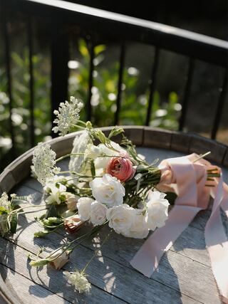 A bouquet of delicate white and soft pink flowers rests on a wooden surface, adorned with a flowing ribbon and surrounded by greenery.
