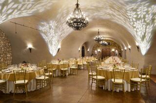 An elegant dining space with gold tablecloths, wooden chairs, and decorative chandeliers, accented by soft lighting on stone walls.