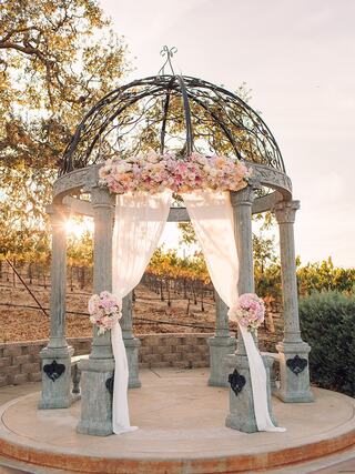 A beautiful gazebo adorned with floral arrangements and draped fabric, set against a serene vineyard backdrop at sunset.