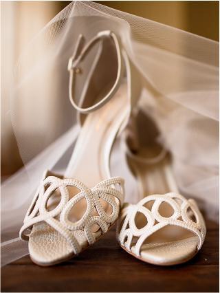 Elegant ivory bridal heels adorned with delicate cut-out designs, resting on a wooden surface partially draped in soft tulle.