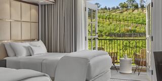 A stylish luxury hotel room with two beds, large windows open to a scenic view of rolling green hills and vineyard, enhancing a serene atmosphere.
