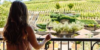 A woman gazes at lush vineyards holding a glass of red wine, surrounded by elegant seating under shaded umbrellas.