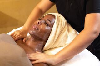 A woman relaxes during a facial treatment, with a towel on her head and a serene expression, as a therapist gently massages her shoulders.