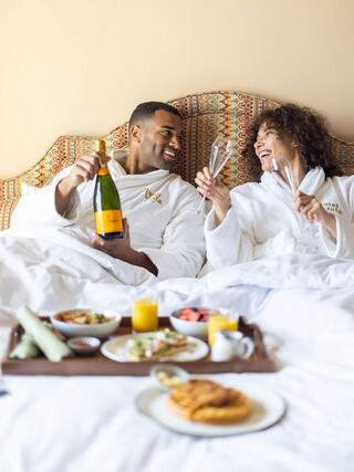 A joyful couple in white robes shares champagne and breakfast in bed, radiating happiness and togetherness.