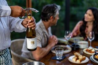 A waiter is pouring wine at a dinner table where two guests enjoy a meal and conversation. Plates of food and drinks are visible.