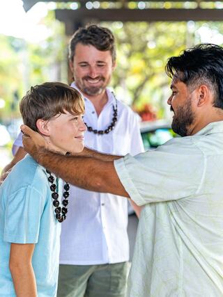 A boy receives a necklace from a man, while another man smiles in the background, all enjoying a warm, friendly atmosphere outdoors.