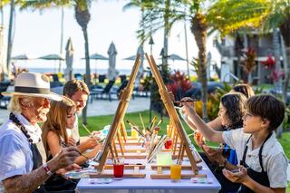A group of people, including children and an older man, enjoy a painting session outdoors, surrounded by palm trees and a beach view.