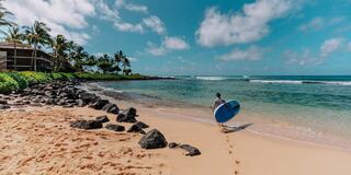 A person walks along a sandy beach, carrying a blue surfboard, with clear water, rocks, and lush greenery under a bright sky.