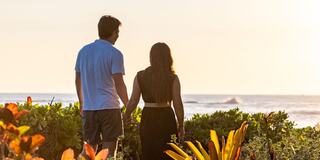 A couple stands hand in hand, gazing at the ocean as the sun sets, surrounded by lush greenery and vibrant flowers.
