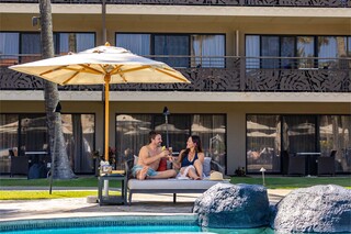 A couple relaxes by a pool, enjoying drinks under an umbrella, surrounded by lush greenery and a stylish hotel backdrop.