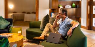 A man relaxes in a cozy lounge, engaged in a phone call while seated comfortably with a laptop nearby. Warm, inviting atmosphere.